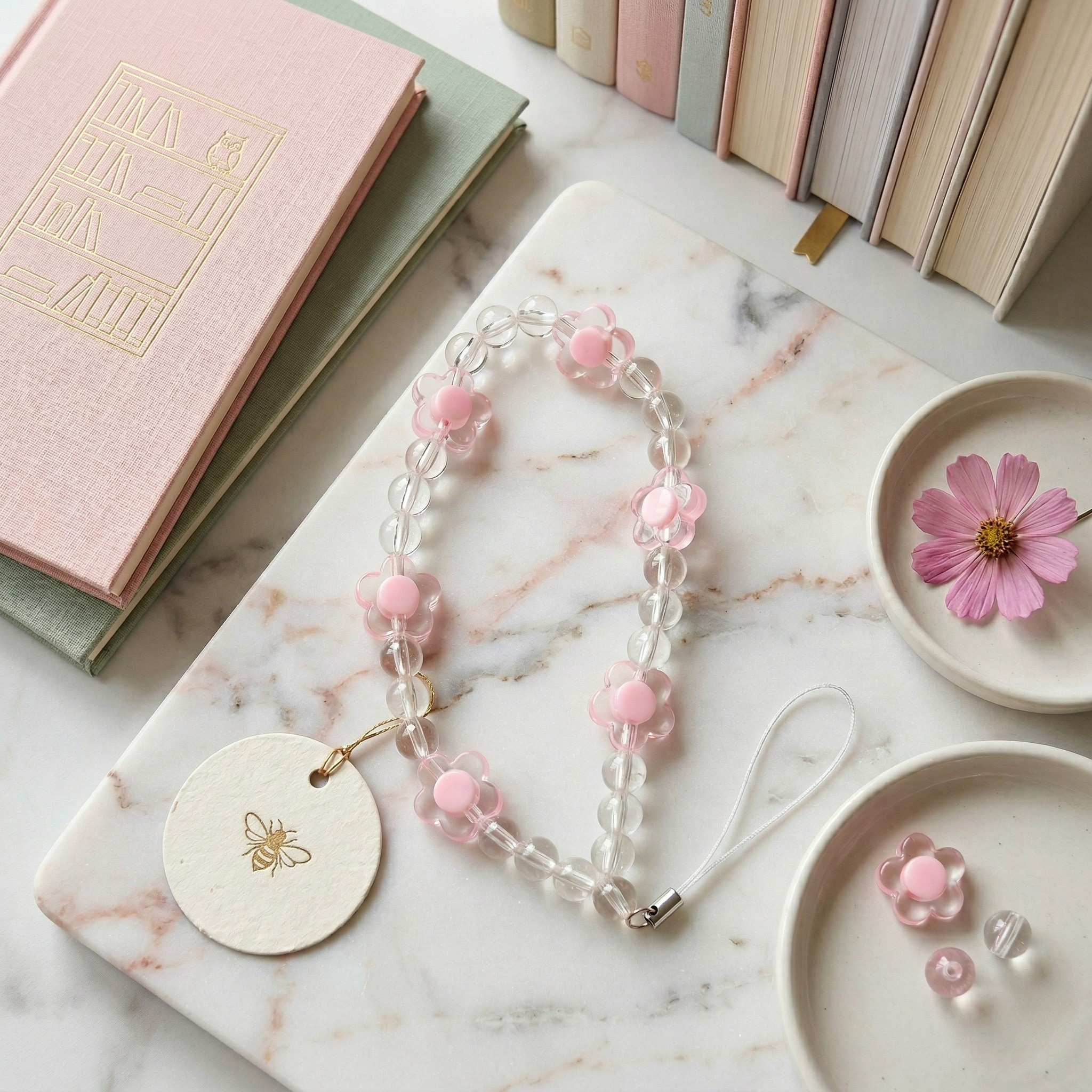 A soft pink and clear glass beaded phone wristlet with flower shapes, styled flat on a marble board alongside pastel books and a small pink cosmos flower.